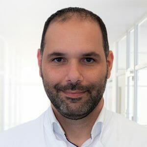 Ein Mann mittleren Alters mit kurzen dunklen Haaren und einem gepflegten Vollbart lächelt freundlich in die Kamera. Er trägt ein weißes Hemd und steht vor einem hellen, unscharfen Hintergrund.