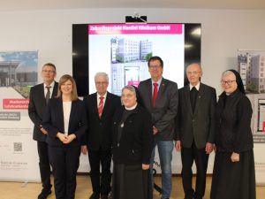 Eine Gruppe von sieben Personen, darunter Klinikvertreter und Ordensschwestern, steht vor einer Präsentation des Zukunftsprojekts der Intensivstation am Nardini Klinikum.