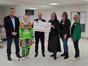 Sechs Personen, darunter eine Ordensschwester und ein Bankvertreter, halten einen symbolischen Spendenscheck über 2.200 Euro für das Nardini Klinikum. Eine Frau trägt einen Alterssimulationsanzug.