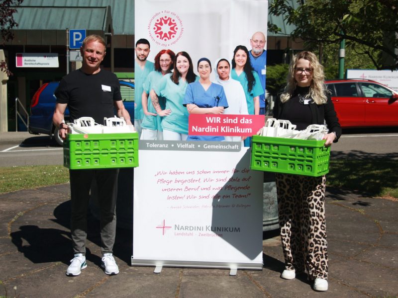 Zwei Personen stehen vor einem Roll-up des Nardini Klinikums und halten grüne Kisten mit kleinen Geschenktüten. Im Hintergrund sind Autos und ein Klinikgebäude zu sehen.