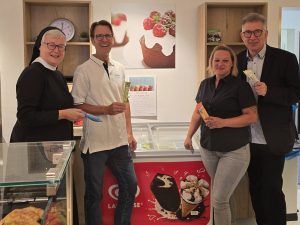Vier Personen, darunter eine Nonne, stehen lächelnd in einem Eiscafé. Sie halten Eis und Geldscheine in den Händen. Im Hintergrund sind Werbeplakate für Eiscreme zu sehen.