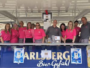 Eine Gruppe von zehn Personen in pinken und grauen T-Shirts steht lächelnd hinter einem Bierstand mit der Aufschrift "Carlsberg". Sie tragen Namensschilder und einheitliche T-Shirts mit einem weißen Sternsymbol. Der Stand befindet sich im Freien bei Tageslicht.