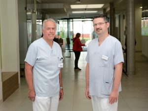 Zwei männliche Ärzte in blauen und weißen Kitteln stehen in einem hell erleuchteten Krankenhausflur. Sie lächeln in die Kamera, während im Hintergrund Personen vorbeigehen.