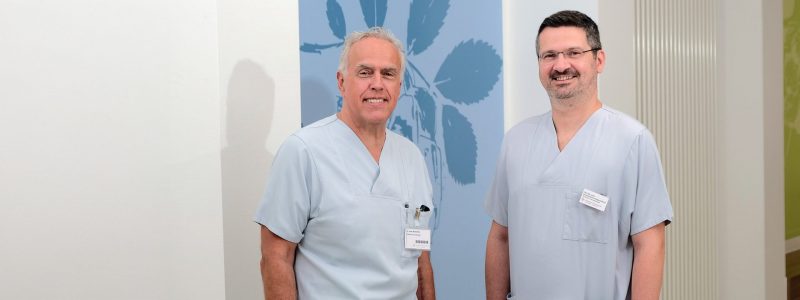 Zwei lächelnde männliche Ärzte in blauen OP-Kitteln stehen in einem Krankenhausflur neben einer Wand mit blauem Blumenmuster.