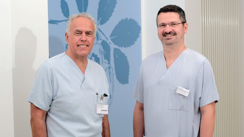 Zwei lächelnde männliche Ärzte in blauen OP-Kitteln stehen vor einer Wand mit einem großen, blauen Blattmuster. Der ältere Arzt links trägt ein Namensschild mit Barcode, der jüngere rechts ein Namensschild mit Text.