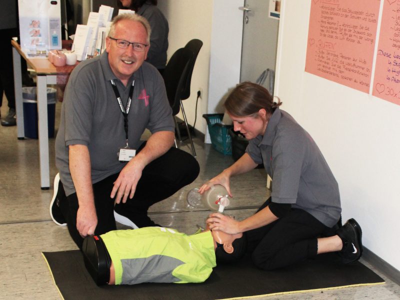 Zwei Personen, eine Frau und ein Mann, demonstrieren Wiederbelebungsmaßnahmen an einer Übungspuppe in einer Schulungsumgebung. Die Frau ist dabei, eine Herzdruckmassage durchzuführen, während der Mann kniend zusieht und lächelt. Im Hintergrund sind Informationsmaterialien auf einem Tisch und Aushänge an der Wand sichtbar.