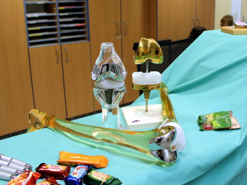 Verschiedene medizinische Geräte, darunter eine goldene und eine silberne Prothese, ausgestellt auf einem türkisfarbenen Tuch in einem Raum mit Holzschränken und Süßigkeiten auf dem Tisch.