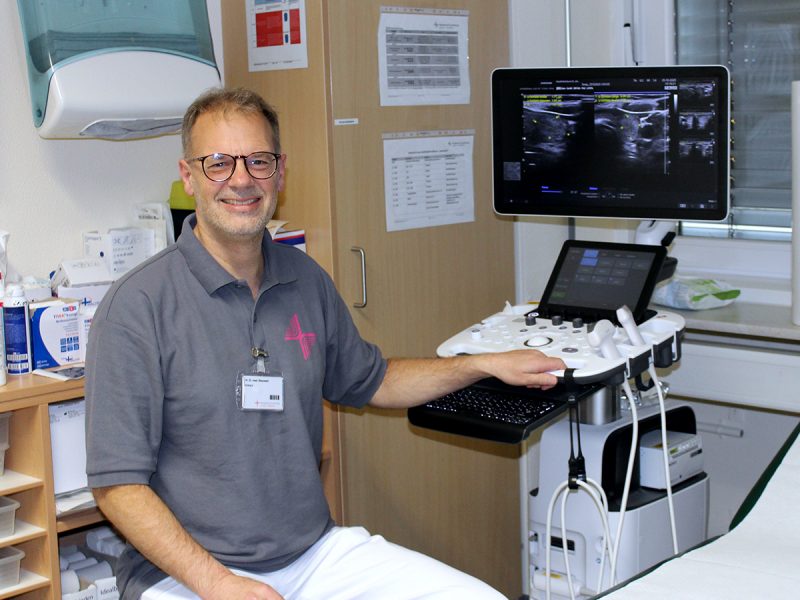 Ein lächelnder Arzt in grauem Poloshirt und weißer Hose sitzt neben einem Ultraschallgerät in einem medizinischen Untersuchungsraum. Auf dem Bildschirm des Geräts sind Ultraschallaufnahmen zu sehen.