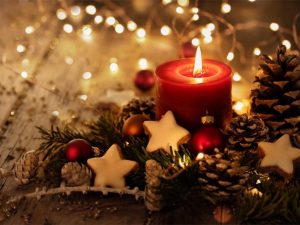 Eine festliche Weihnachtsdekoration mit einer brennenden roten Kerze, umgeben von Tannenzweigen, Kiefernzapfen, roten Kugeln und Sternplätzchen, alles beleuchtet von sanftem Lichterkettenschimmer auf einem Holztisch.