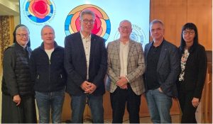 Sechs Personen stehen nebeneinander vor einem farbenfrohen Glasfenster mit Rosenmotiv während einer Veranstaltung.