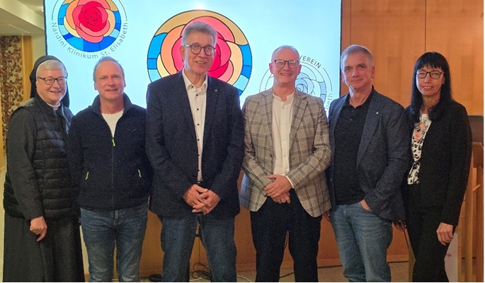 Sechs Personen stehen nebeneinander vor einem farbenfrohen Glasfenster mit Rosenmotiv während einer Veranstaltung.