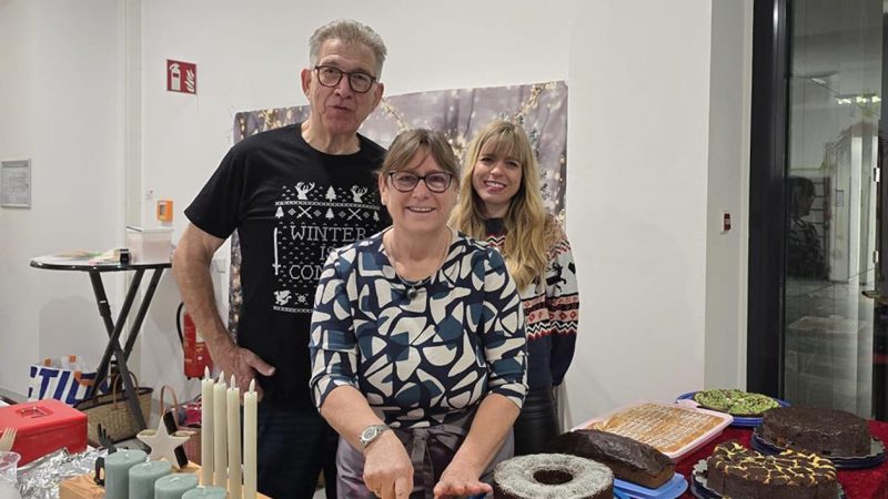 Drei Personen stehen hinter einem Tisch mit verschiedenen Kuchen bei einer Veranstaltung. Ein Mann und zwei Frauen lächeln in die Kamera. Der Mann trägt ein schwarz-weißes Winter-T-Shirt, die Frauen tragen gemusterte Pullover.