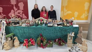 Drei Frauen stehen hinter einem Verkaufsstand in einer hellen Halle, der mit verschiedenen weihnachtlichen Handwerksartikeln dekoriert ist. Auf dem Tisch liegen unter anderem handgemachte Krippen, Weihnachtssterne und Adventskränze auf einem türkisfarbenen Tuch. Im Hintergrund sind große, abstrakte Silhouetten in Rot und Gelb zu sehen.