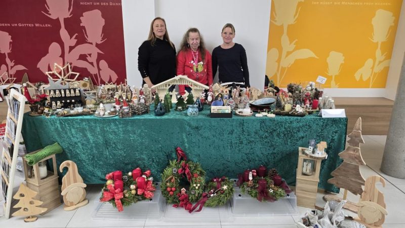 Drei Frauen stehen hinter einem Verkaufsstand in einer hellen Halle, der mit verschiedenen weihnachtlichen Handwerksartikeln dekoriert ist. Auf dem Tisch liegen unter anderem handgemachte Krippen, Weihnachtssterne und Adventskränze auf einem türkisfarbenen Tuch. Im Hintergrund sind große, abstrakte Silhouetten in Rot und Gelb zu sehen.