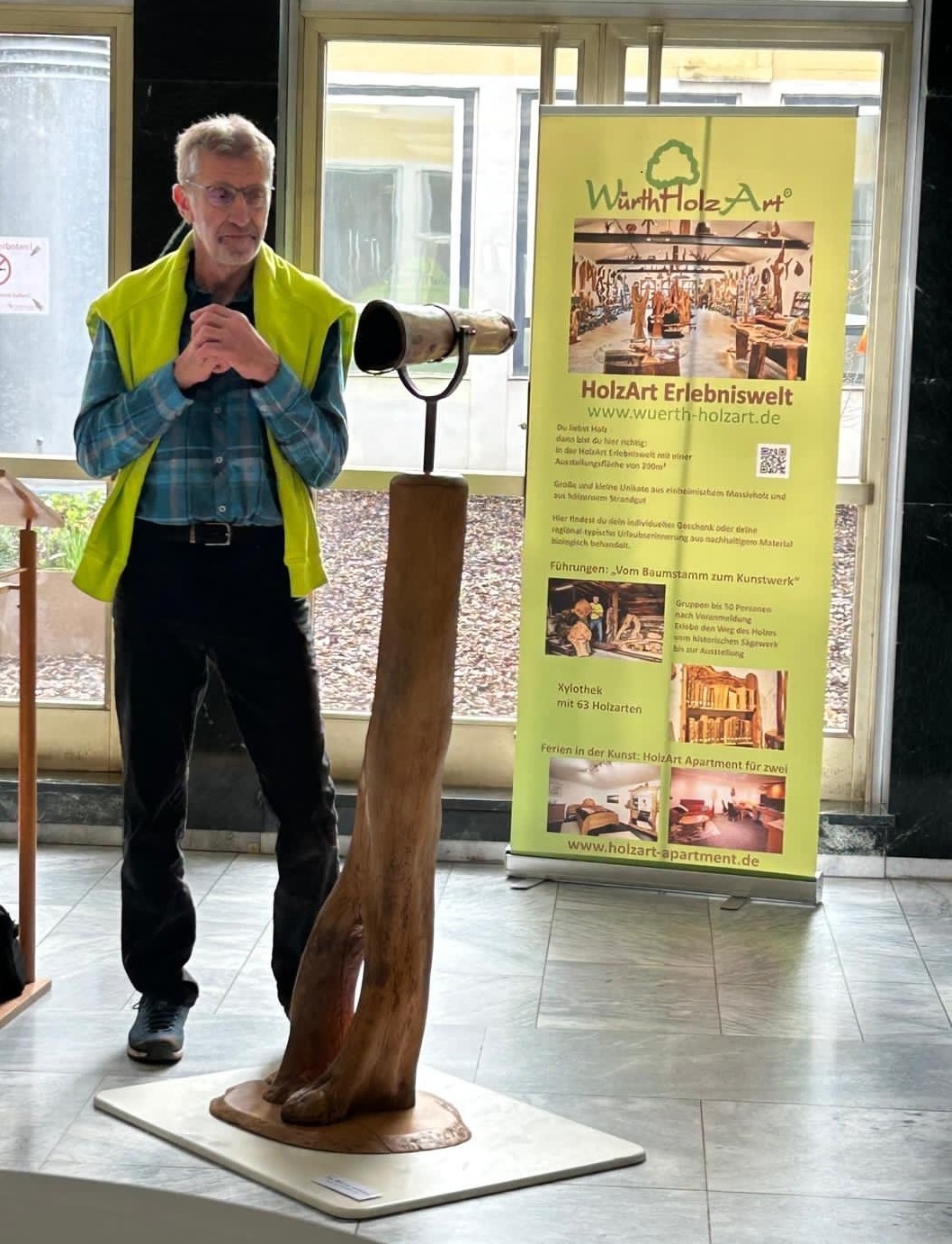Ein älterer Mann spricht vor einer Holzskulptur in der Eingangshalle des Nardini Klinikums. Im Hintergrund steht ein Werbebanner von Würth HolzArt.