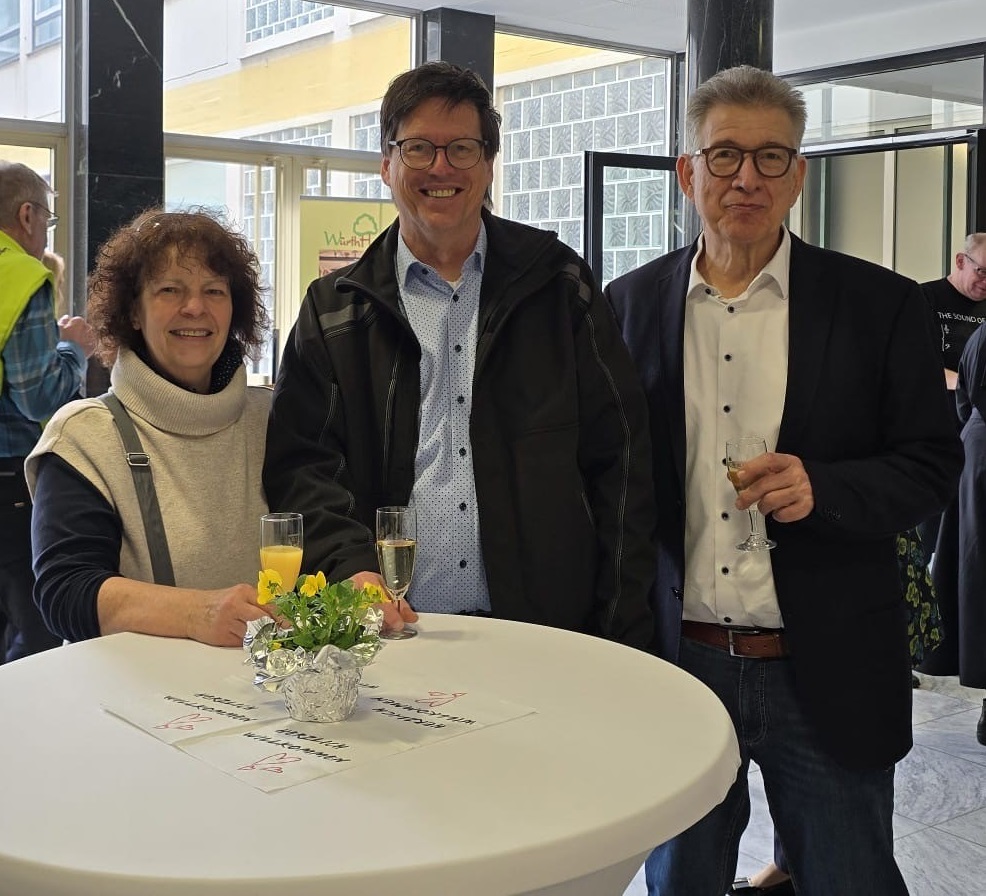 Drei Personen stehen an einem Stehtisch mit Getränken, umgeben von Dekoration.